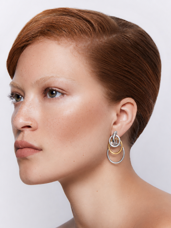A close-up side profile of a woman with short auburn hair wearing a pair of stacked, interlocking hoop earrings in silver and gold tones against a light gray background. 